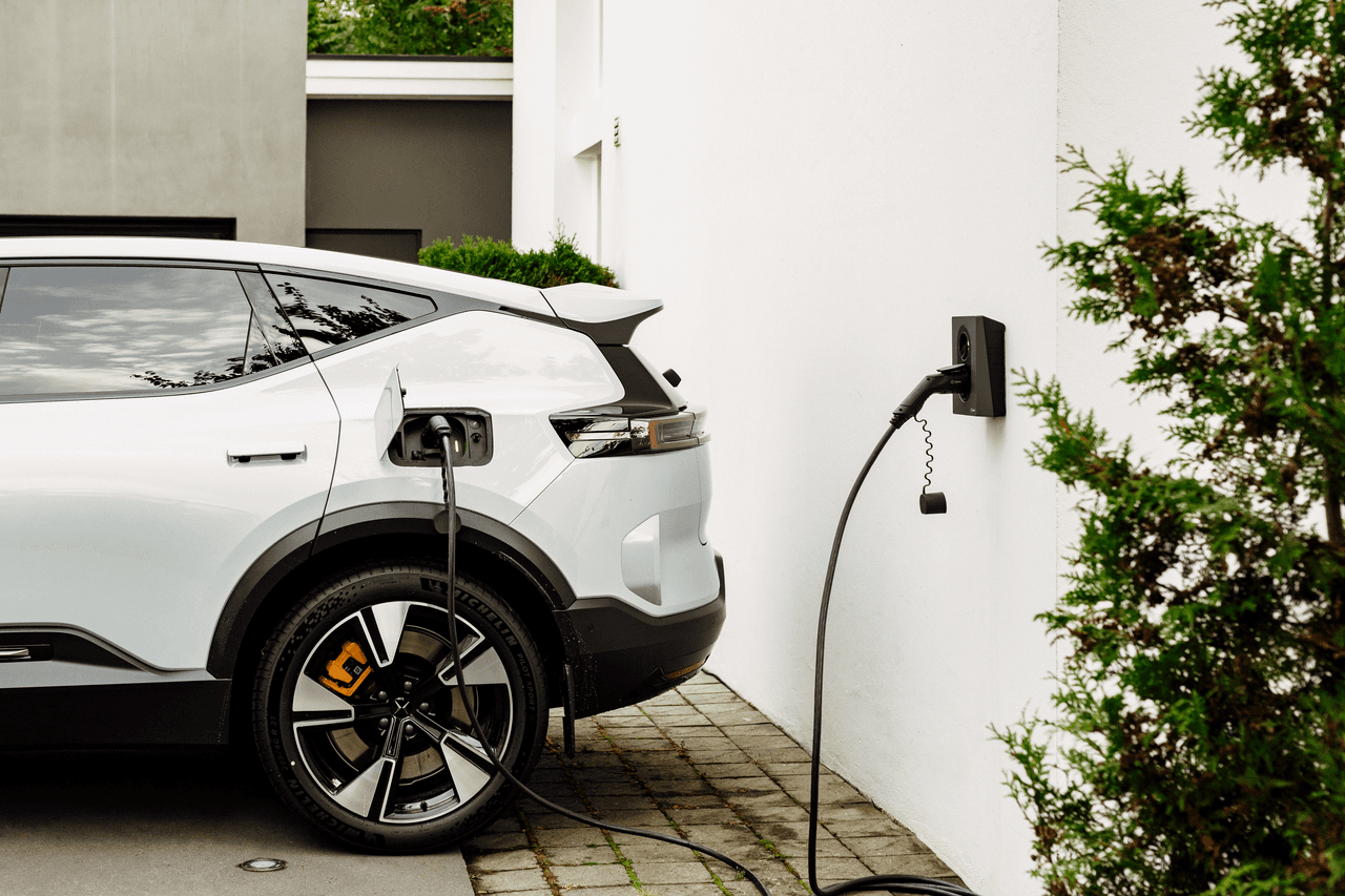 En hvit elbil lades med en svart Zaptec Go 2-lader som er montert på en hvit husvegg, omgitt av grøntområder.