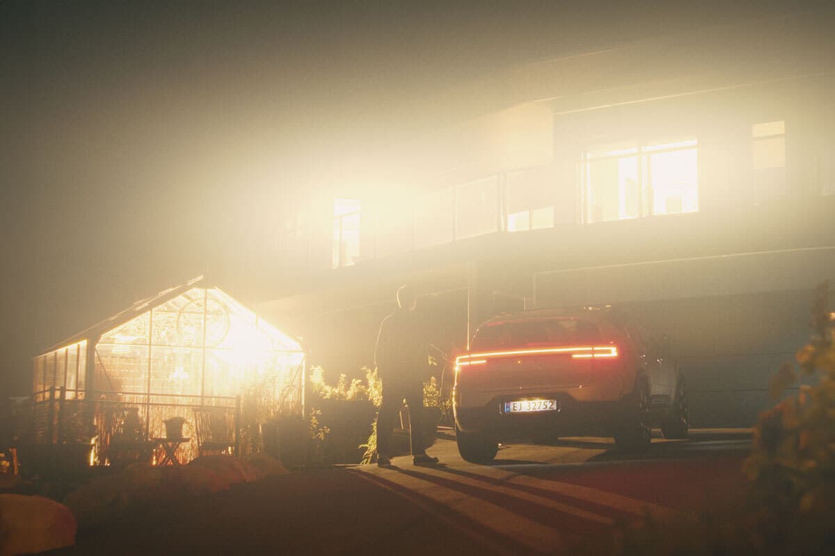 Nattbilde av en oppkjørsel foran en moderne bolig. En elbil står med baklyktene tent, og en person står ved siden av bilen. Et glassdrivhus og huset lyser kraftig opp.