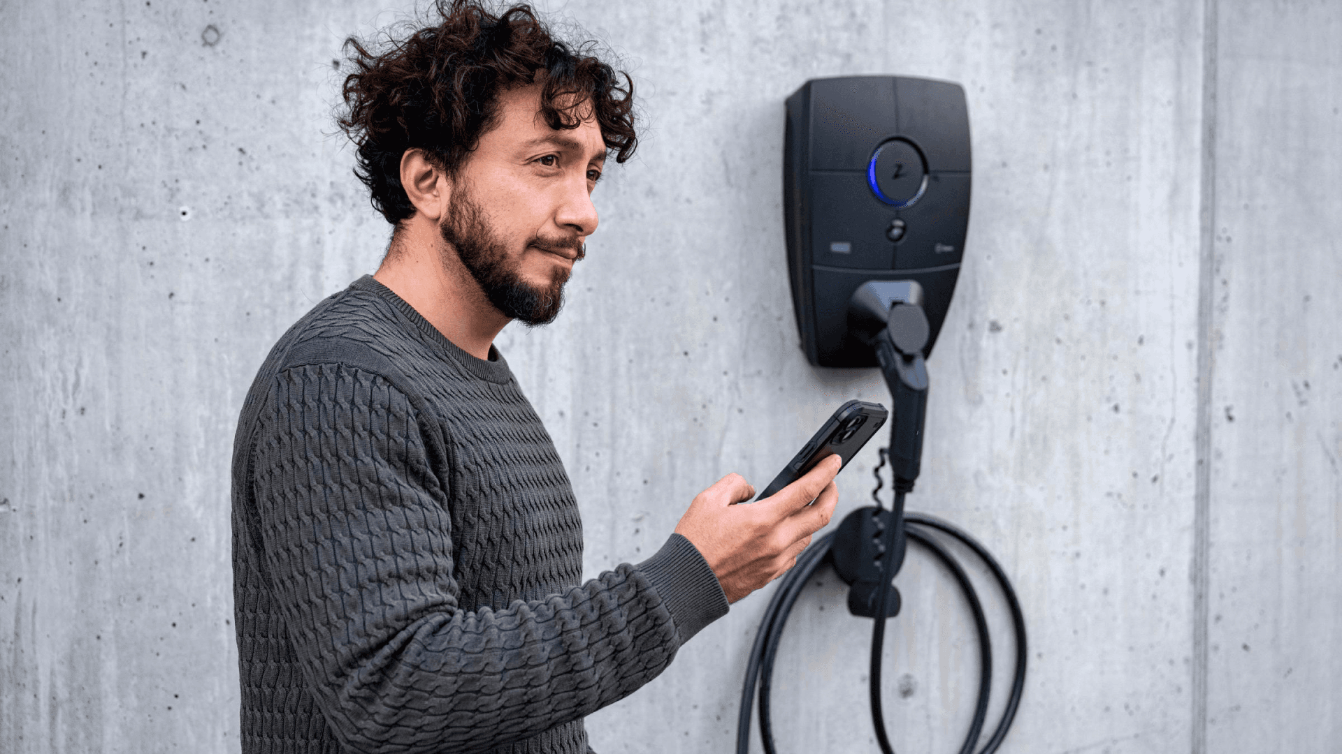 En mann med krøllete hår og grå genser står foran en Zaptec Pro-lader montert på en betongvegg. Han holder en mobiltelefon og ser på skjermen mens ladekabelen henger pent oppkveilet.