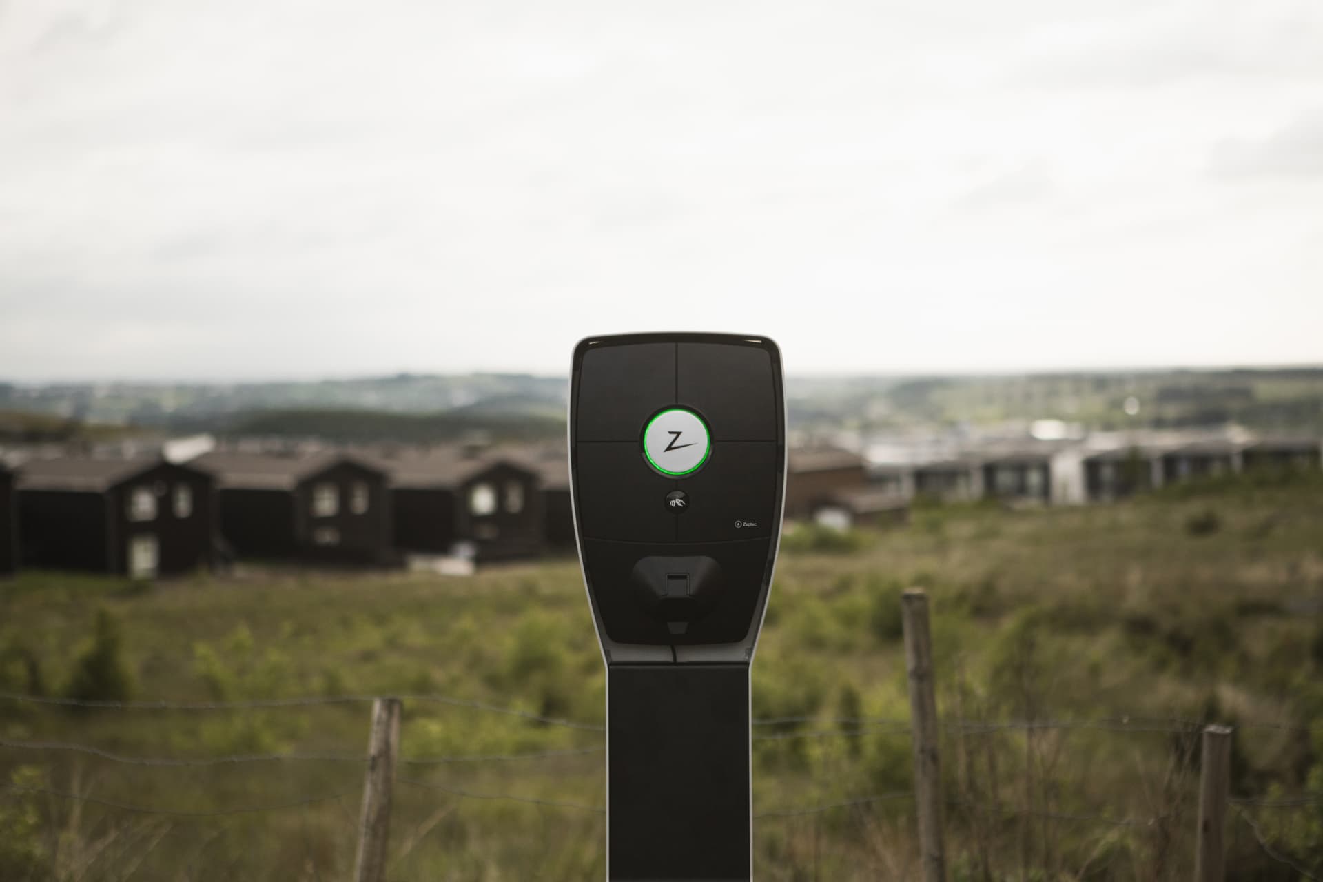 En Zaptec Pro-lader er montert på en svart stolpe i et gresskledd område med moderne trehus og et vidstrakt landskap i bakgrunnen.