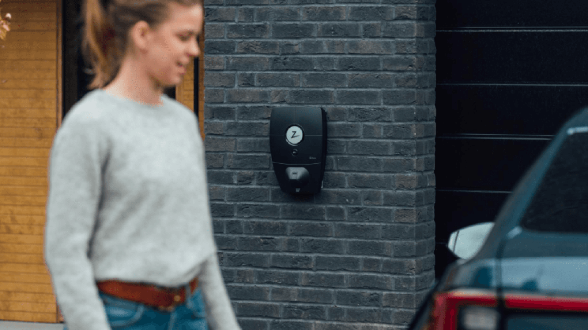 En kvinne i en grå genser går forbi en Zaptec Pro-lader montert på en mørk murvegg. En bil er delvis synlig i forgrunnen.