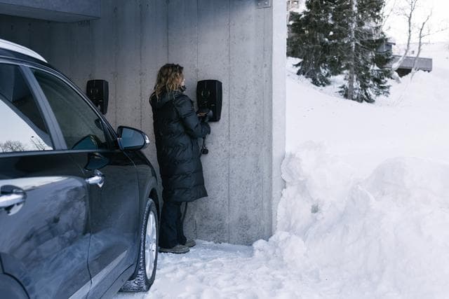 En kvinna iklädd en lång svart vinterjacka kopplar in en laddkabel i en Zaptec Pro laddare monterad på en betongvägg i ett snöigt garage. Bredvid henne står en svart bil parkerad.