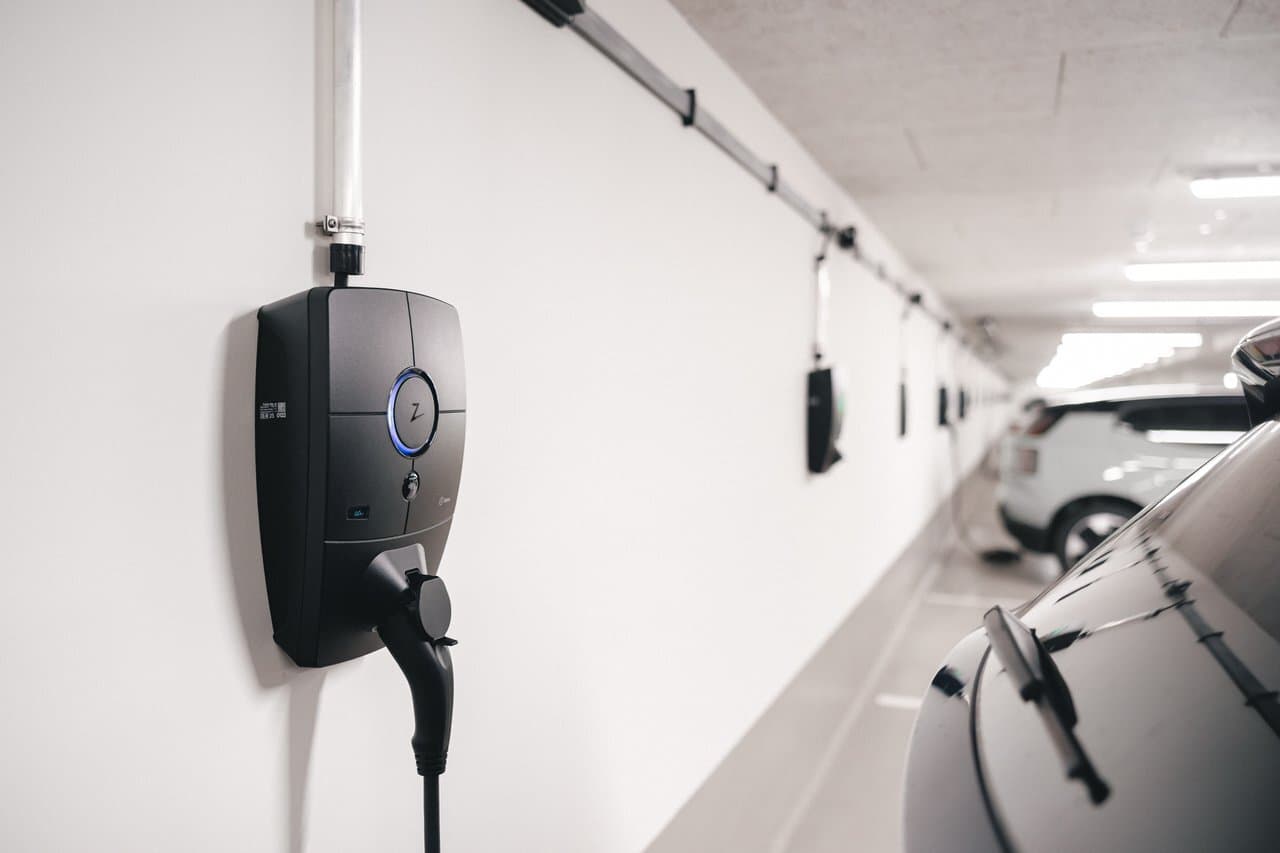 En rekke Zaptec Pro-ladere er montert langs en hvit vegg i et innendørs parkeringshus. Flere elbiler er tilkoblet og lader.