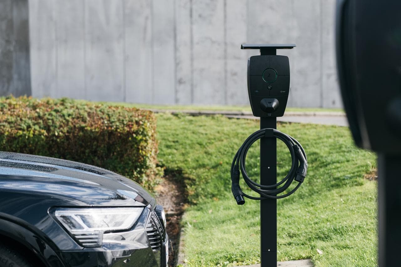 En svart bil er parkert ved siden av en Zaptec Pro-lader montert på en svart stolpe i et grønt uteområde med klipte hekker og en betongvegg i bakgrunnen.