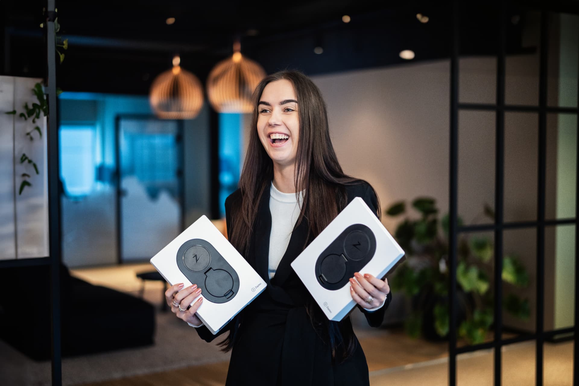 En smilende kvinne i mørke klær holder to Zaptec Go-ladere i fargen Cloud White i et moderne kontormiljø.