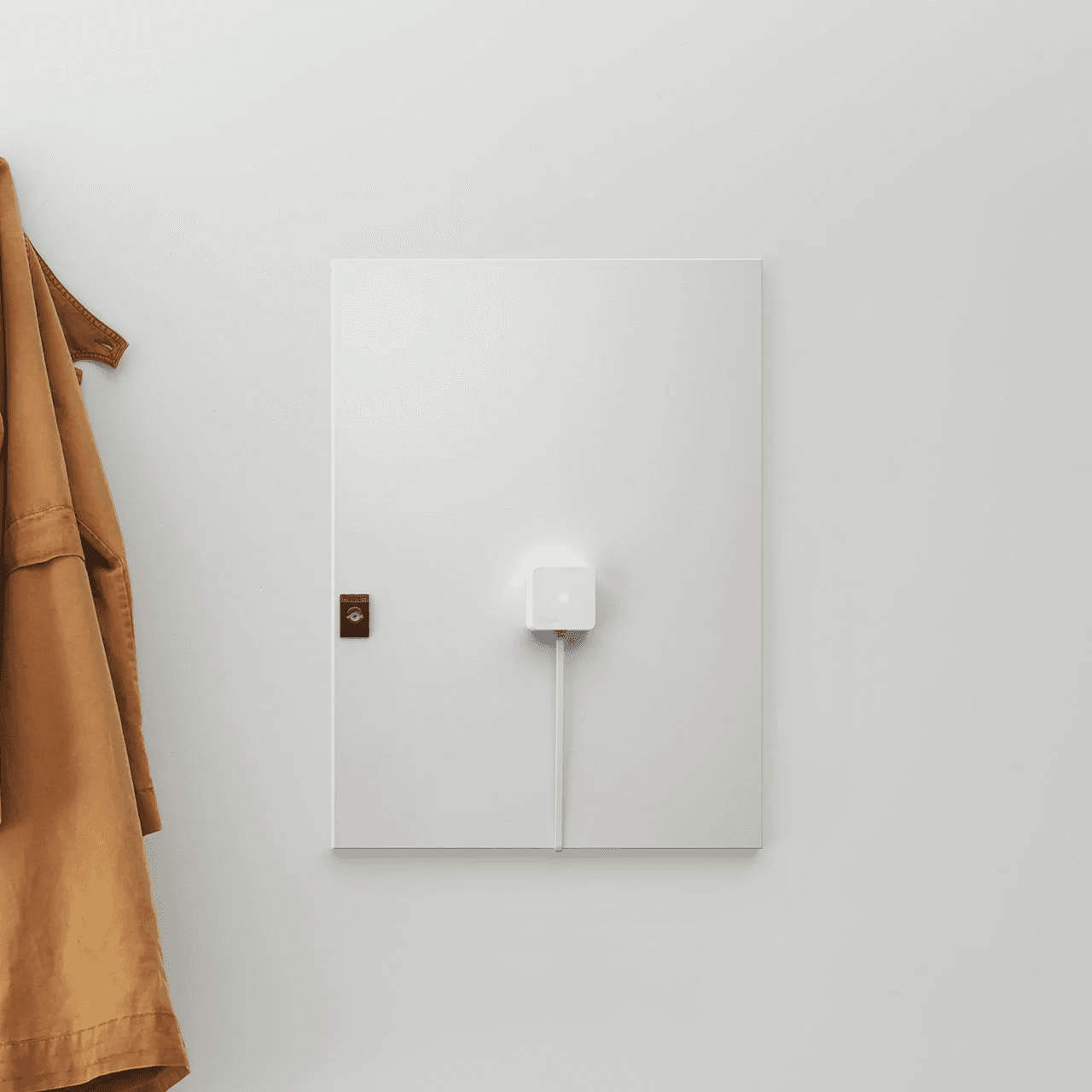 En hvit Zaptec Sense er montert på et skap i en lys gang med en brun jakke hengende ved siden av. Enheten er koblet til med en hvit kabel.