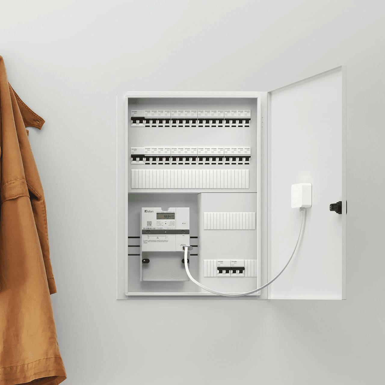 Et åpent sikringsskap med en installert Zaptec Sense koblet til strømmåleren med en hvit kabel. En brun jakke henger ved siden av på en lys vegg.