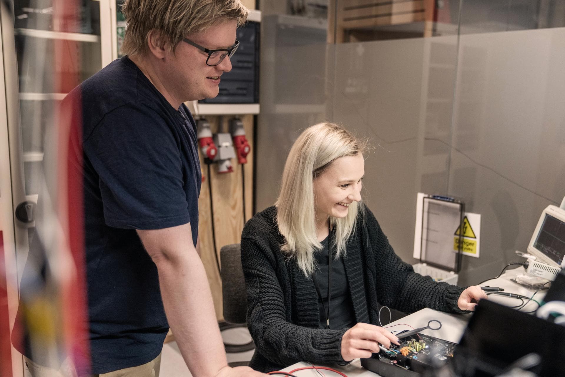 To kollegaer jobber sammen ved en elektronikkarbeidsstasjon. Kvinnen sitter og smiler mens hun jobber med et kretskort, og mannen står ved siden av og observerer.