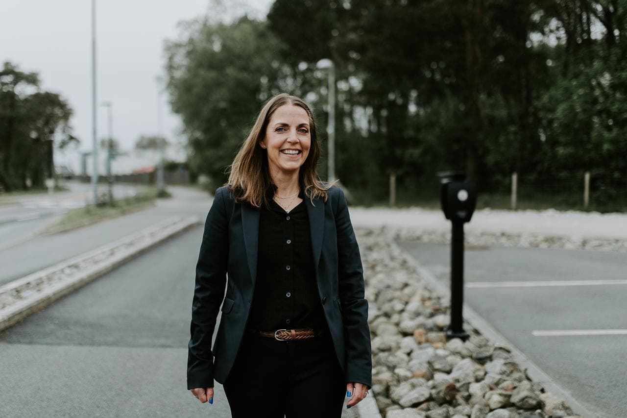 En kvinne i mørk blazer går smilende langs en vei med en Zaptec Pro-lader synlig i bakgrunnen ved en parkeringsplass.