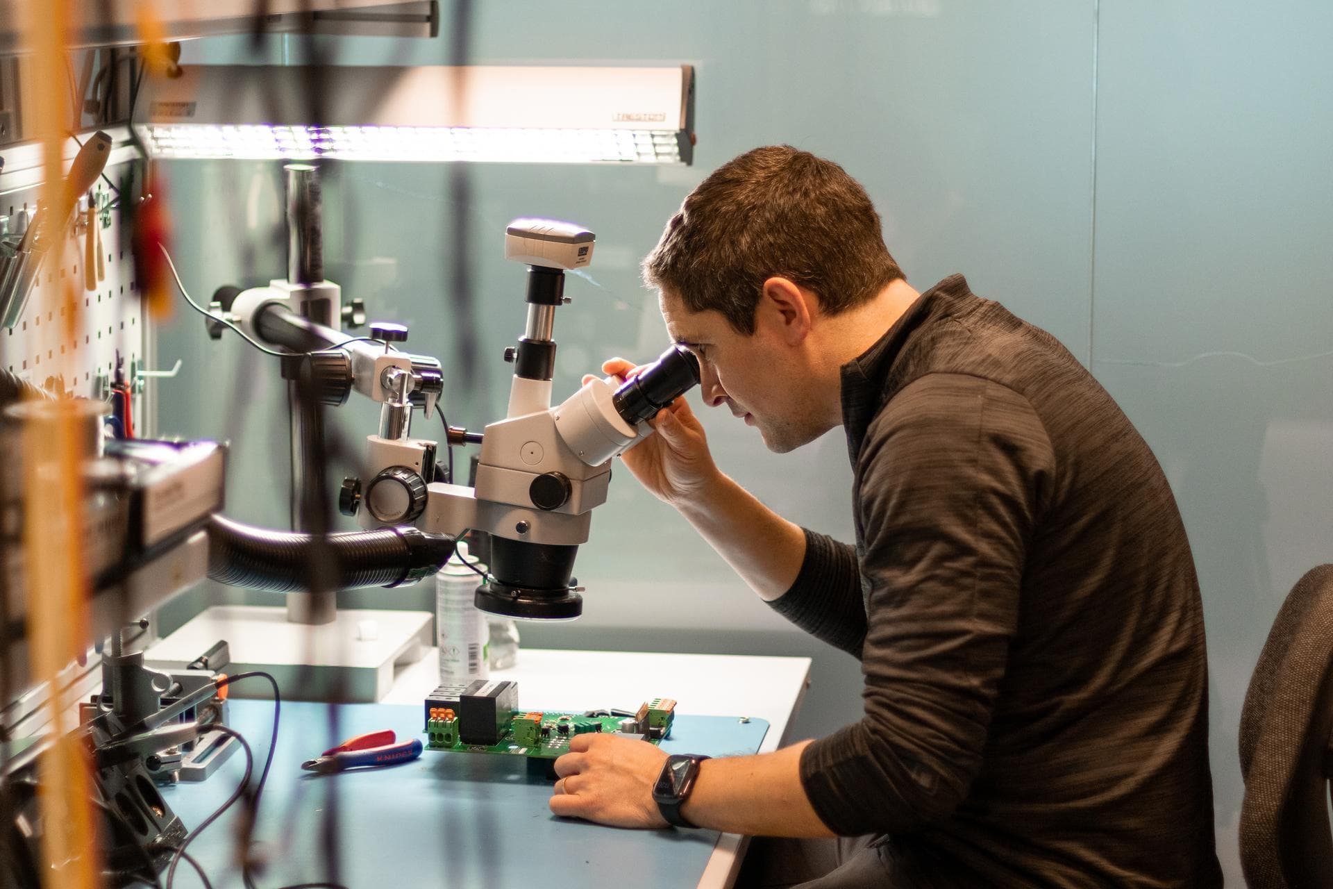En mann undersøker elektroniske komponenter gjennom et mikroskop ved en godt opplyst arbeidsbenk