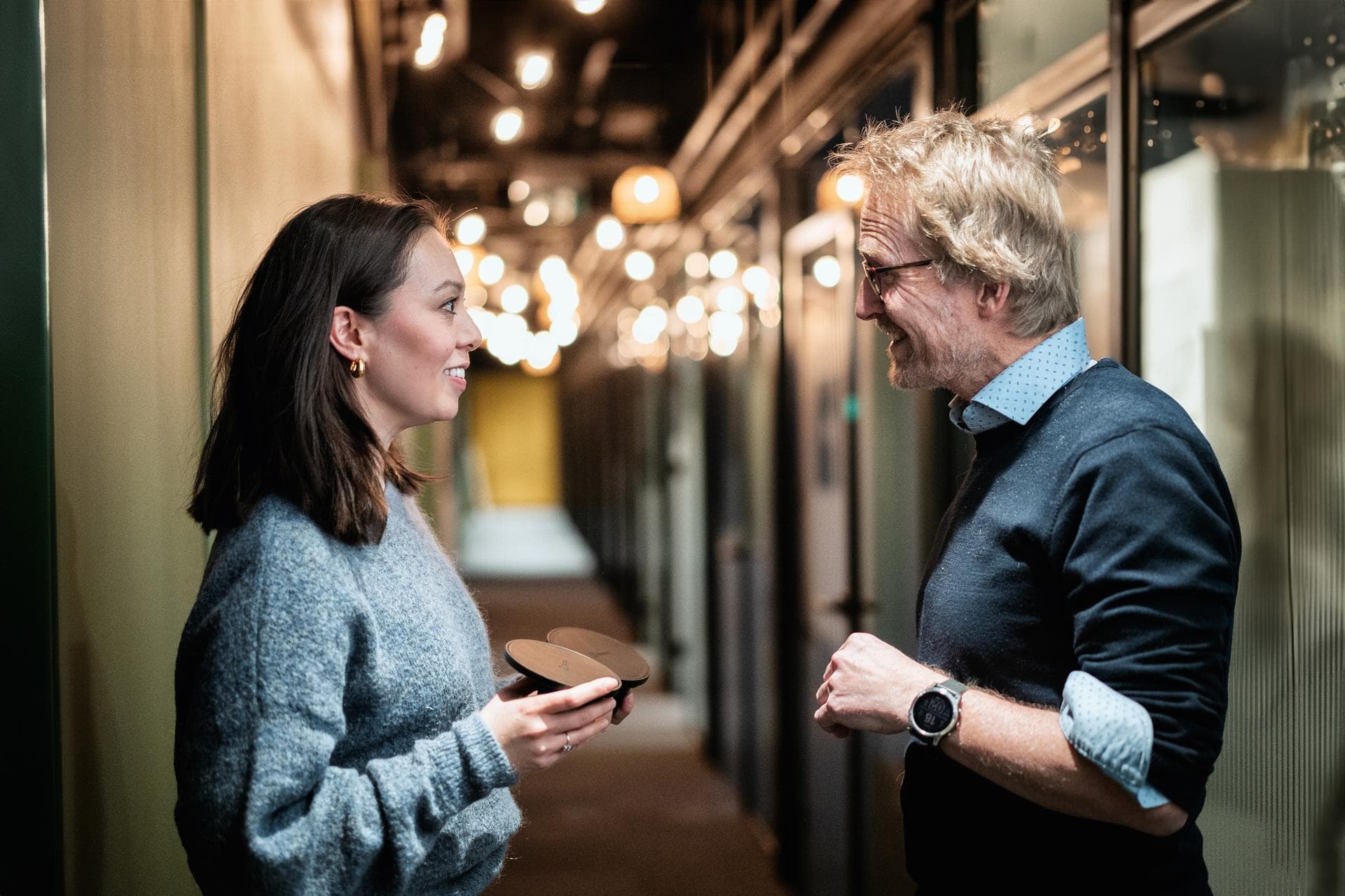 To kollegaer har en vennlig samtale i en varmt opplyst gang. Kvinnen holder en Zaptec Chill og smiler, mens mannen gestikulerer mens han snakker.