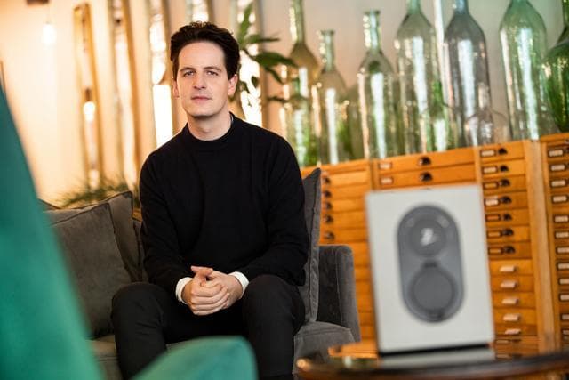 Daniel Gwercher VD Zaptec Tyskland sitter i en nätt fotölj med mörka kläder och mörkt hår med händerna ihopflätade och armbågarna lutade mot låren. I förgrunden syns en Zaptec Go. i bakgrunden syns en mänk med många långsmala lådor och stora silverfärgade flaskor.