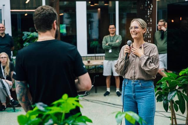 En kvinne holder en mikrofon og smiler mens hun snakker foran en liten gruppe kollegaer på et Zaptec-arrangement. Hun står blant grønne planter og skaper en vennlig og avslappet stemning.