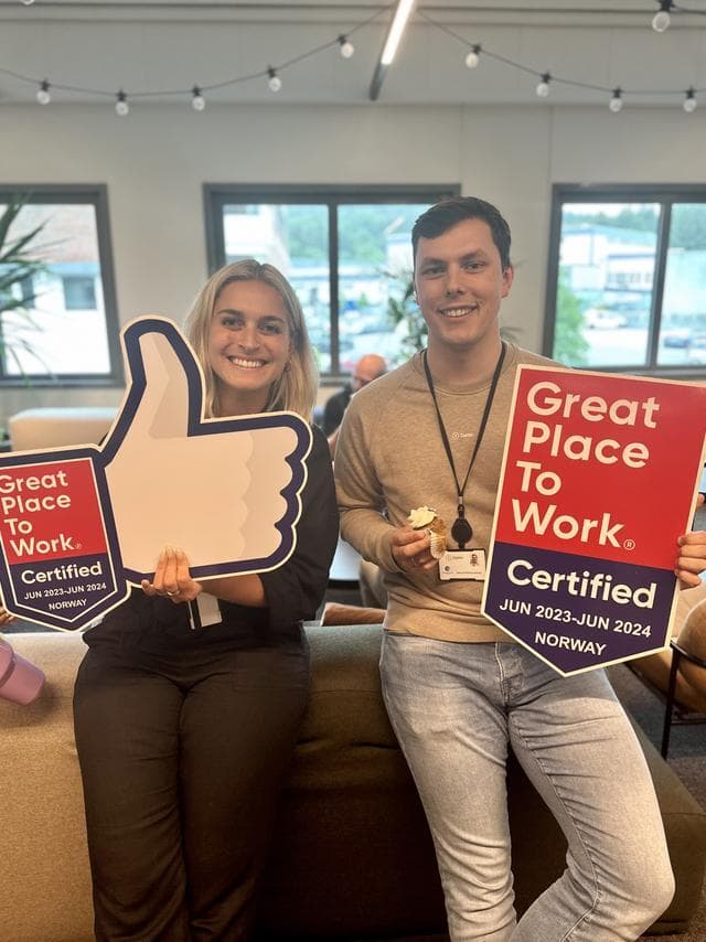 To smilende kollegaer holder opp “Great Place to Work”-skilt i et moderne kontormiljø. Den ene holder en tommel opp-form, den andre et sertifiseringsskilt – en feiring av en positiv arbeidskultur.