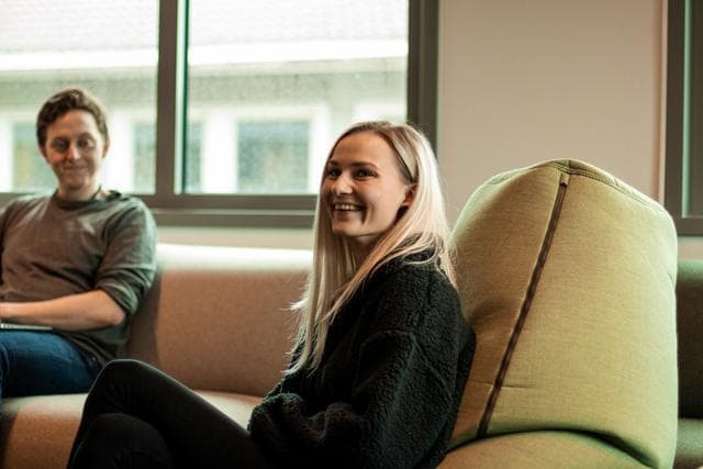 i en grön sittmöbel sitter en glad ljushårig kvinna i mörka kläder, i bakgrunden syns en ljus soffa där det sitter en man och leer han har mörkbrunt kort hår, grön tröja och blå jeans
