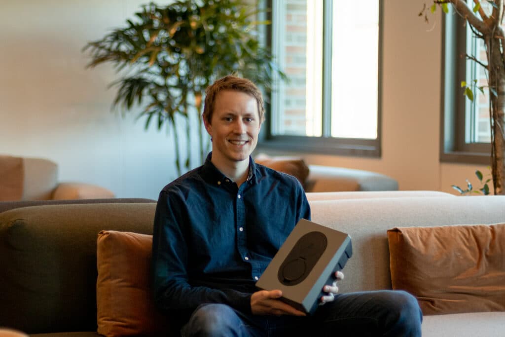 Een glimlachende man in een donkerblauw overhemd zit op een moderne en comfortabele bank. Hij houdt een donkere Zaptec Go-laadpaal in zijn handen. Op de achtergrond laten grote ramen natuurlijk licht binnen, met groene planten en kussens in warme tinten.