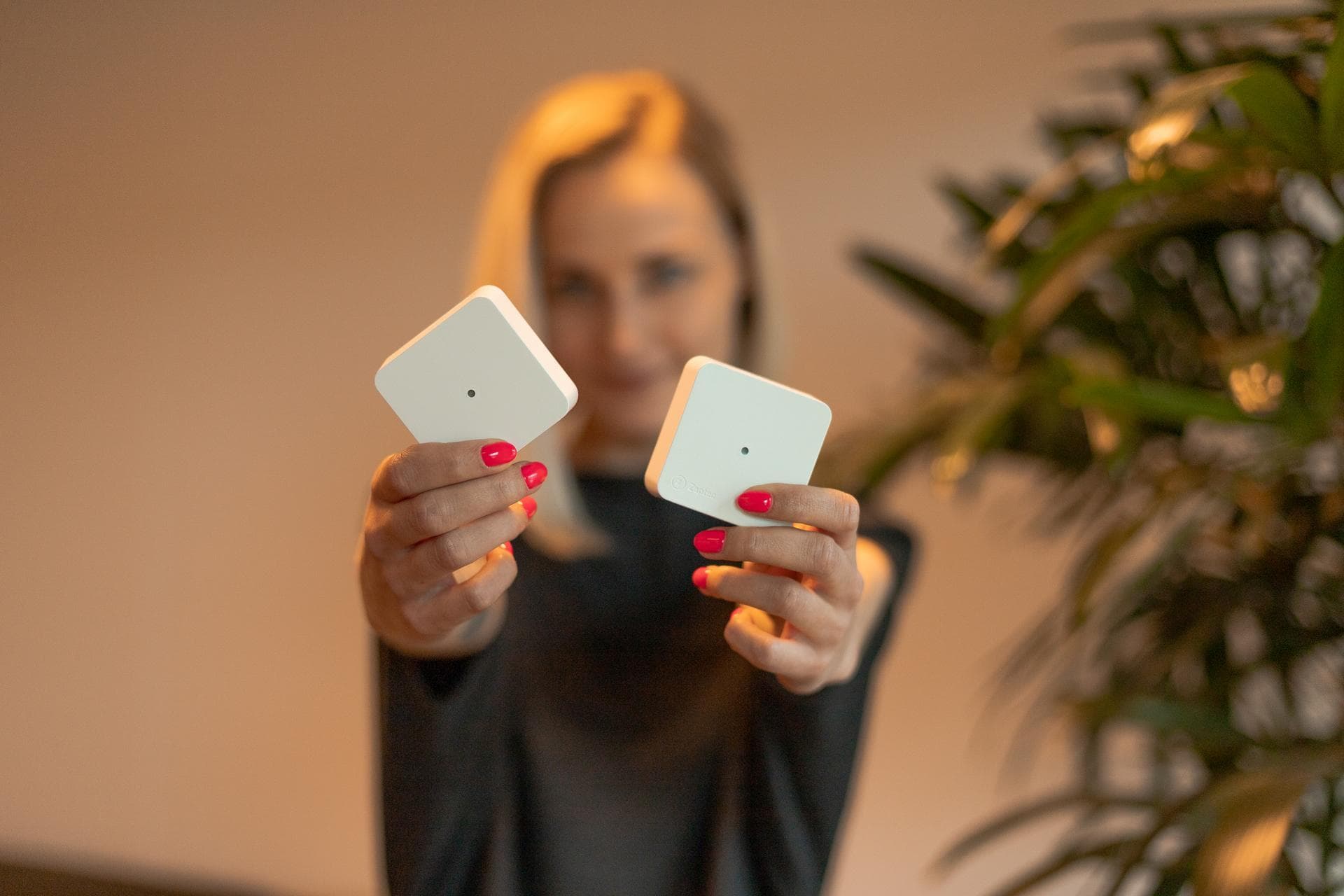En kvinne holder opp to hvite Zaptec Sense-enheter med begge hender, rettet mot kameraet. Hun smiler, og i bakgrunnen sees en grønn plante i mykt lys.