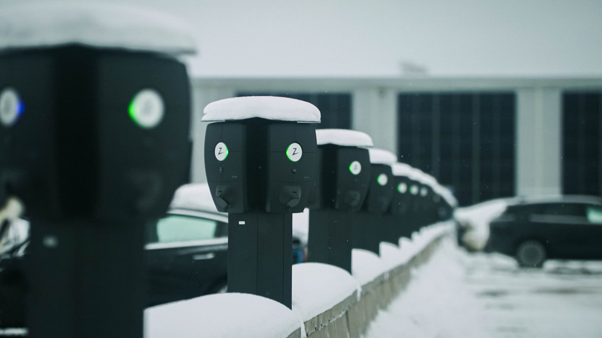En rekke Zaptec Pro-ladere dekket av snø står på rad på en parkeringsplass i vintermiljø, med biler parkert i bakgrunnen.