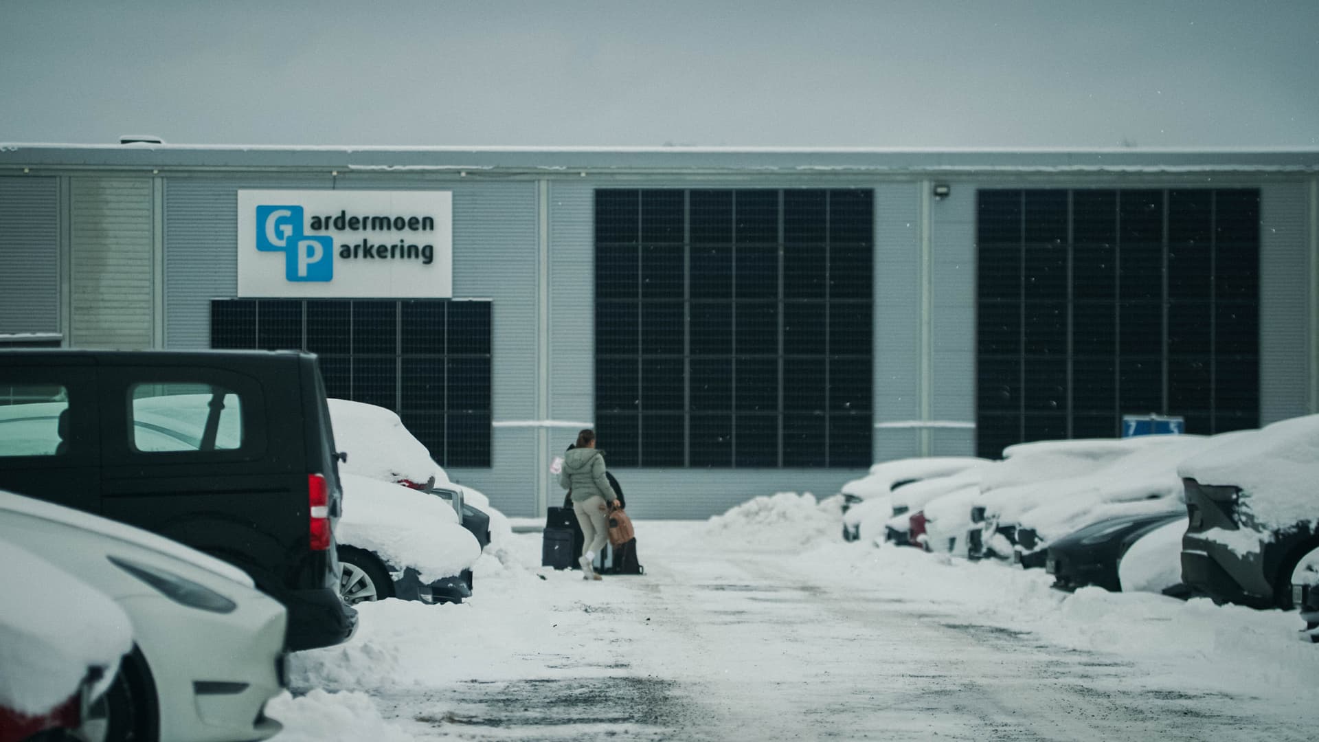 en parkering med snöinbäddade bilar och två personer som drar resväskor i den plogade vägen mellan bilarna