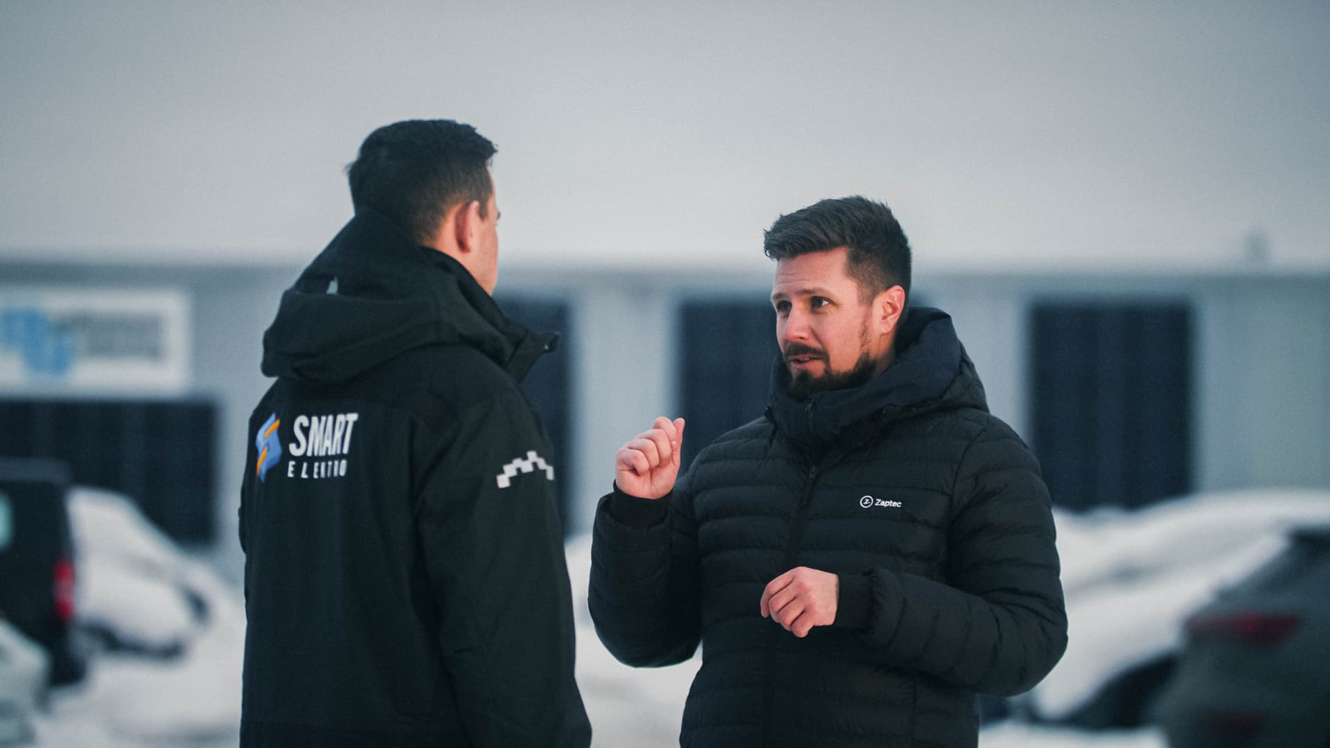 två män med mörka kläder och mörkt hår står och pratar på en snötäckt parkering