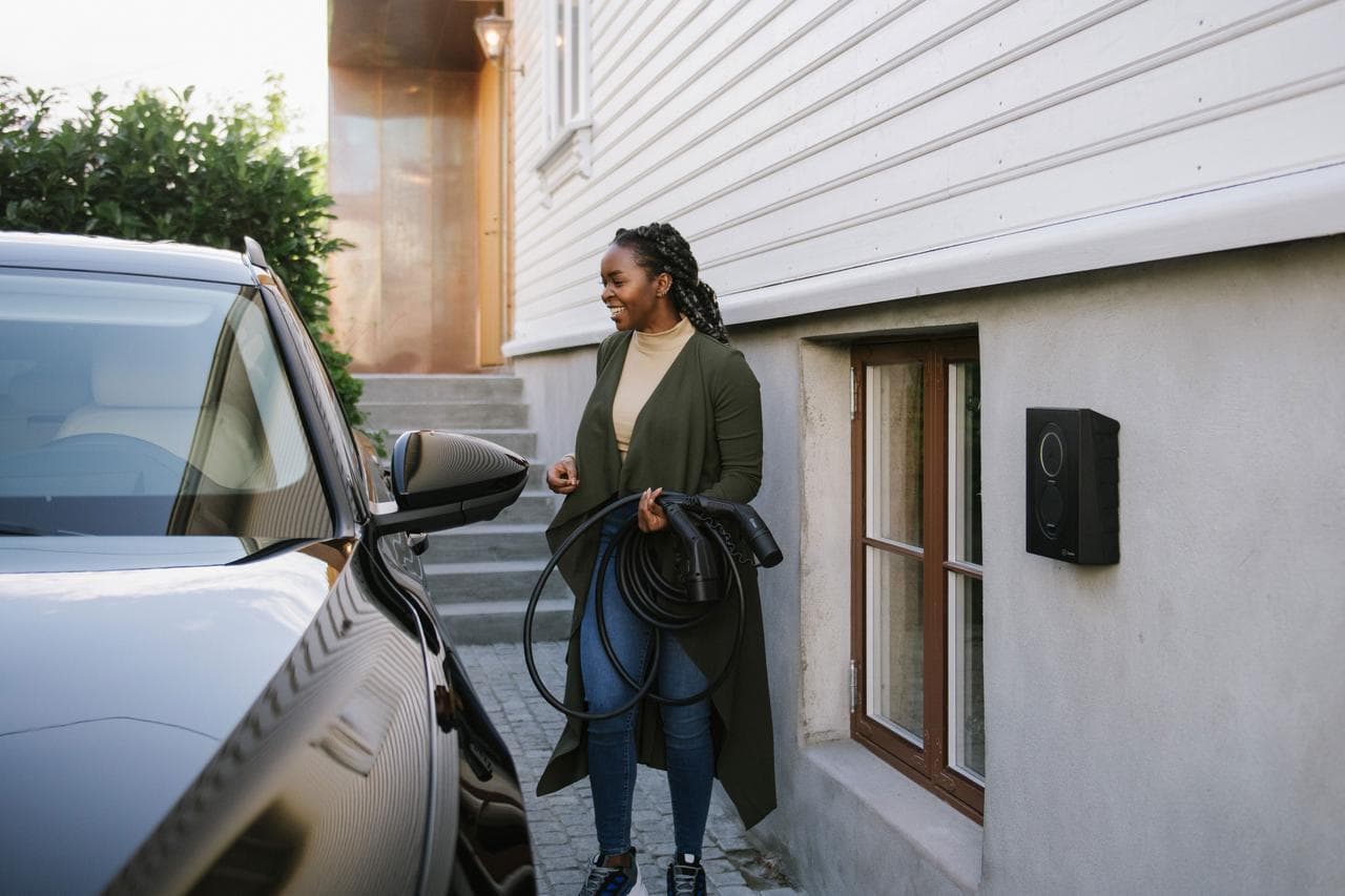 En kvinna står vid en svart elbil som står parketad intill ett hus med vit och grå fasad. Kvinnan håller i en ihopfullad laddkabel och på väggen intill henne sitter en svart Zaptec Go