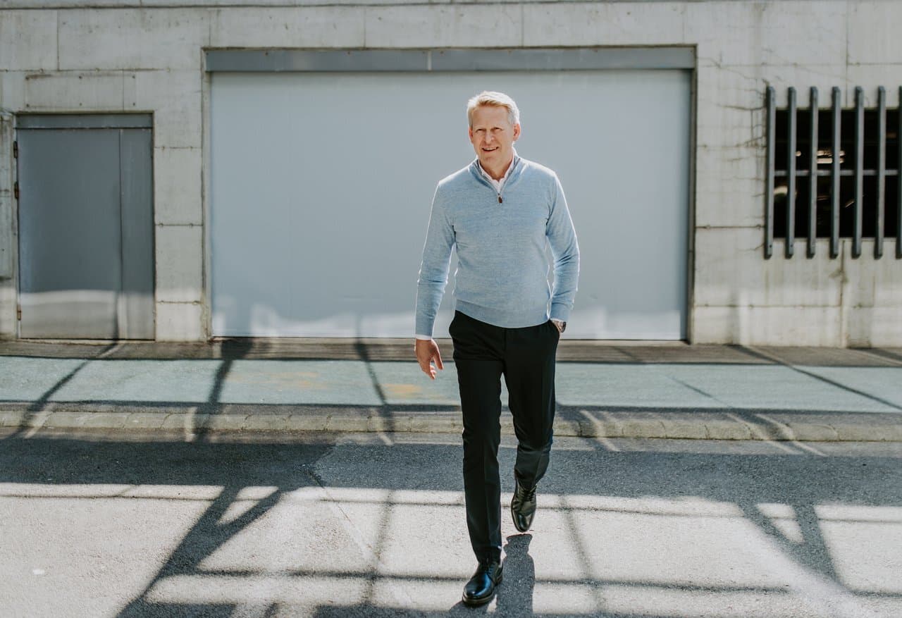 A man with short hair, dressed in a light blue sweater and black trousers, walks confidently outdoors in bright sunlight. He is in front of a large concrete wall with a closed garage door and metal details casting shadows on the ground.
