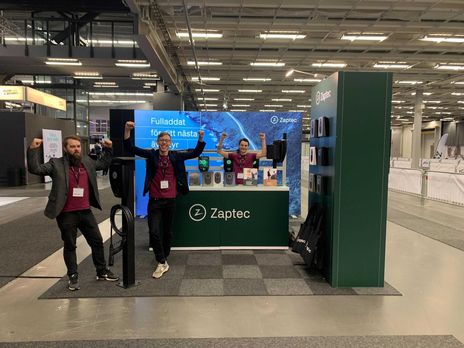 Tre entusiastiske teammedlemmer poserer ved en Zaptec-stand på en messe. Standen viser ladere og utstillingsenheter med grønn profilering, og en skjerm med teksten «Fulladet for ditt neste eventyr».