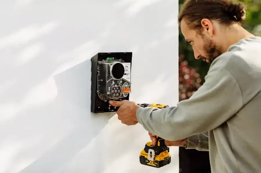 En man i grå tröja installerar en Zaptec-laddare på en vit vägg med hjälp av en gul och svart skruvdragare. Laddarens front är borttagen.