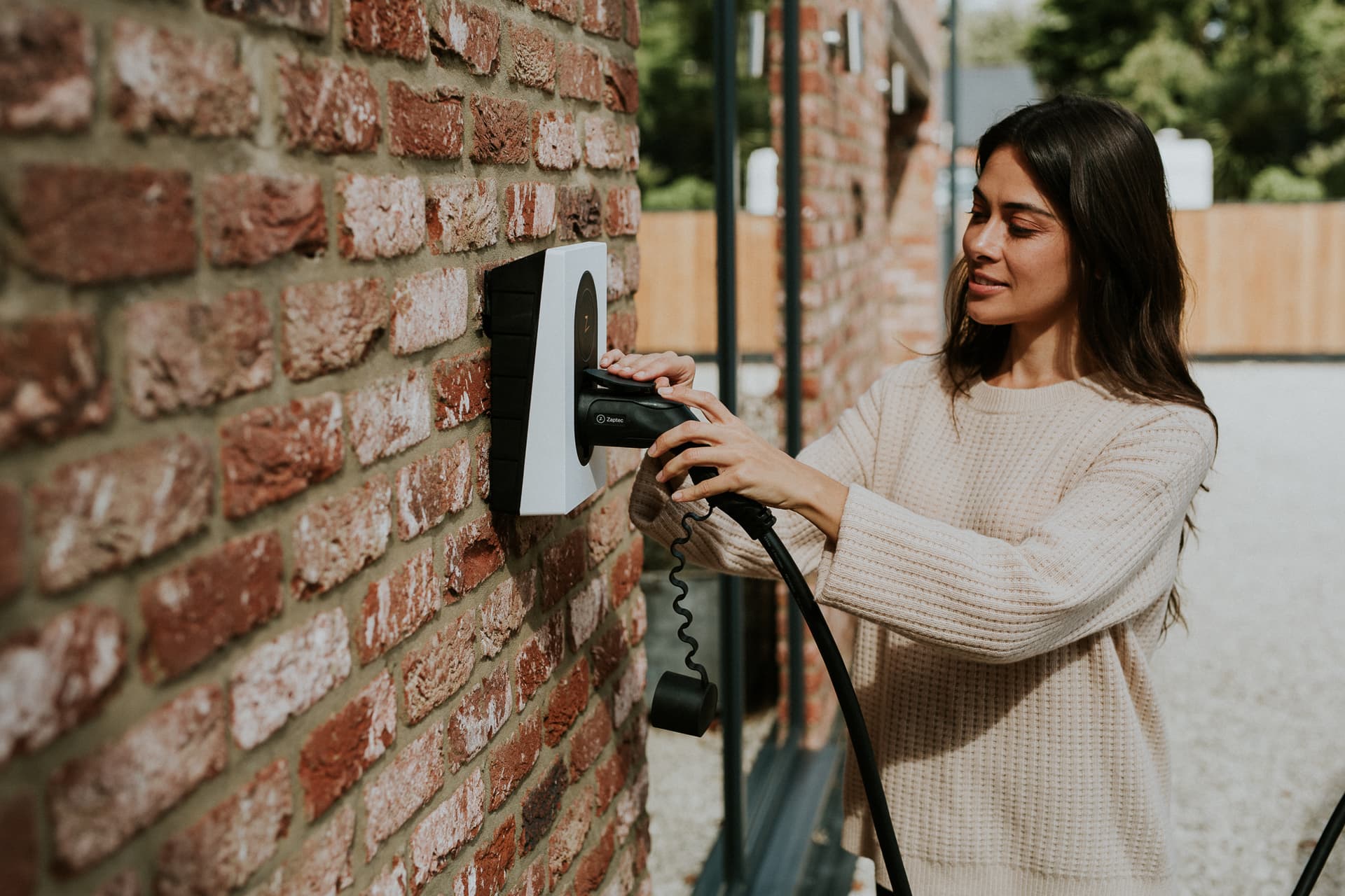 Een vrouw in een beige trui sluit een zwarte laadkabel aan op een witte Zaptec-laadpaal die gemonteerd is op een roodbruine bakstenen muur. De zon schijnt en verlicht de warme tinten van de muur en haar gezicht.